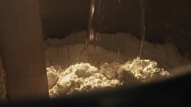 Close-up shot shows water stream falling into flour inside large mixing bowl baker preparing dough for baking process creation of food products at commercial bakery. Baker pours iced water to flour in