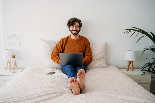 Happy young freelancer sitting on bed with laptop