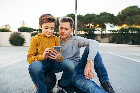 Father and son sitting on basketball outdoor court using cell phone