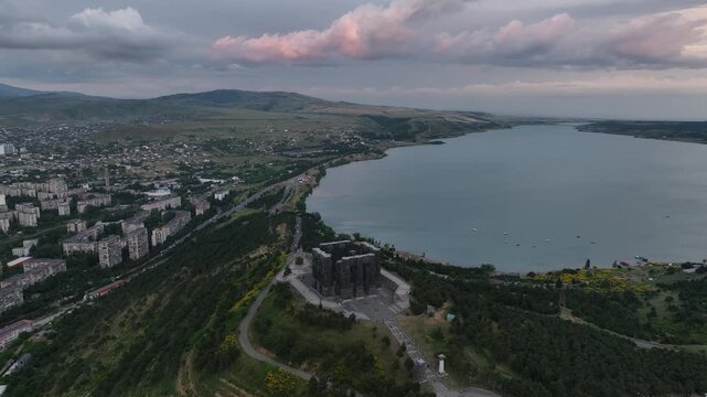 Drone shot capturing the historical Chronicle of Georgia monument complex during a scenic evening sunset. The huge stone pillars are set on a green hill next to the calm Tbilisi Sea reservoir