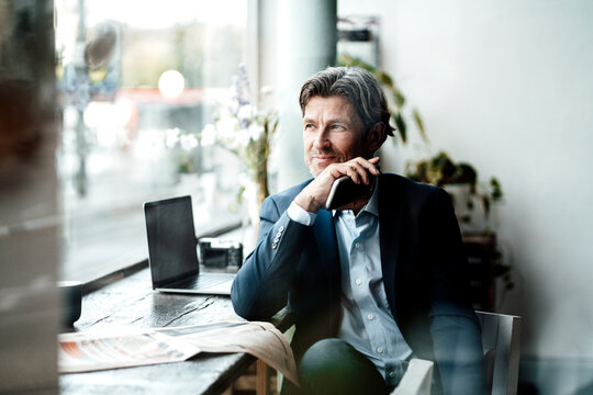 Smiling businessman holding smart phone looking through window while sitting at table in cafe