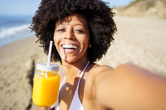 Happy woman taking selfie with orange juice at beach in summer