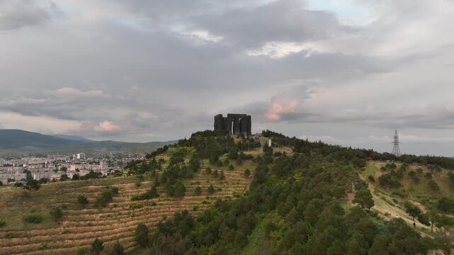 Drone shot capturing the historical Chronicle of Georgia monument complex during a scenic evening sunset. The huge stone pillars are set on a green hill next to the calm Tbilisi Sea reservoir