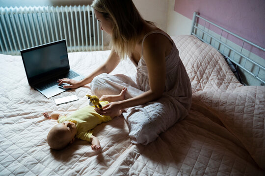 Working mother using laptop and playing with baby boy lying on bed at home