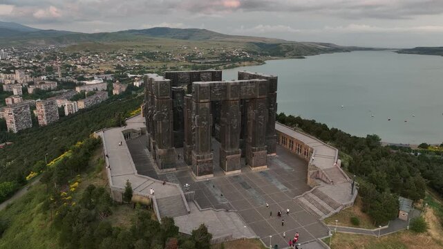 Drone shot capturing the historical Chronicle of Georgia monument complex during a scenic evening sunset. The huge stone pillars are set on a green hill next to the calm Tbilisi Sea reservoir