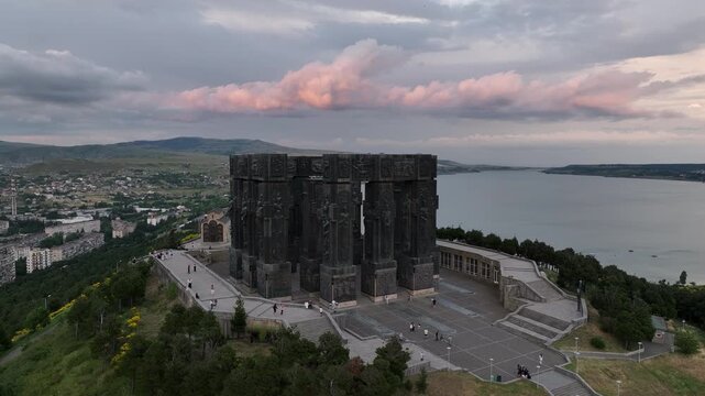 Drone shot capturing the historical Chronicle of Georgia monument complex during a scenic evening sunset. The huge stone pillars are set on a green hill next to the calm Tbilisi Sea reservoir