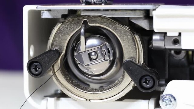 Detailed view of sewing machine lower thread system, featuring metal bobbin case, shuttle hook, and bobbin with black thread. Intricate mechanical process for fabric stitching and textile production.
