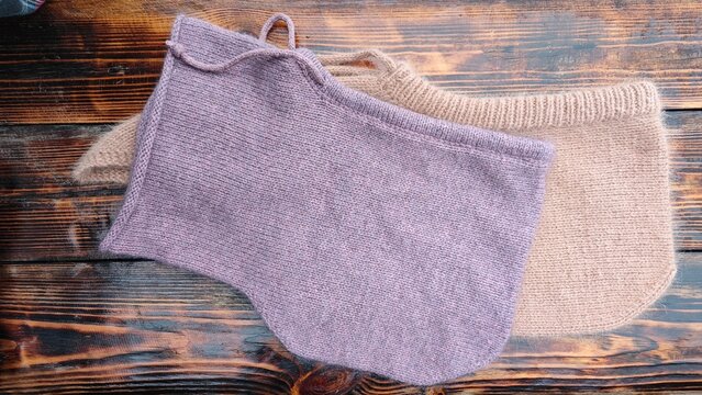Horizontal flat lay of two handmade knitted winter hoods on a dark rustic wooden table. Dusty pink and beige wool balaclavas. Cozy winter fashion accessories