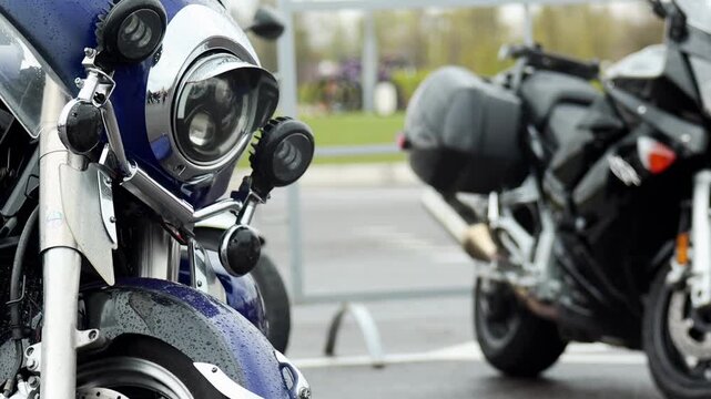 Close-up perspective of blue motorcycle headlight and front section, detailed view of chrome elements, wet surface with raindrops visible on paint and windshield, parked vehicle at outdoor event.