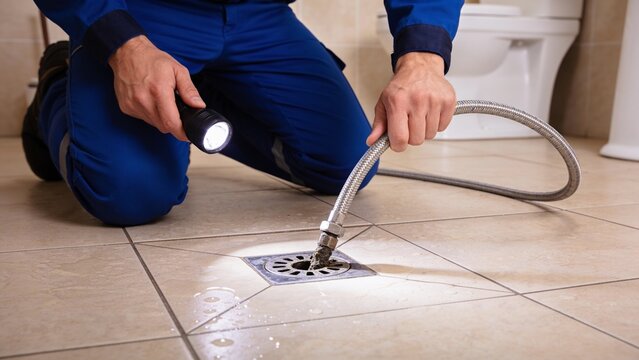 A plumber in blue workwear kneeling to inspect a clogged floor drain with a flashlight and a hose.