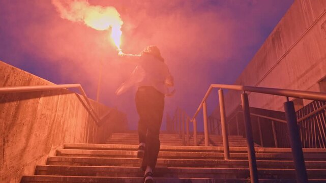 A skateboarder in casual attire leaps down a staircase at dusk, a flare casting a vivid glow, exuding daring and youthful exuberance.