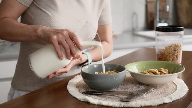 Woman pours fresh milk from a pitcher into a cereal bowl on kitchen counter. Prepares morning breakfast.