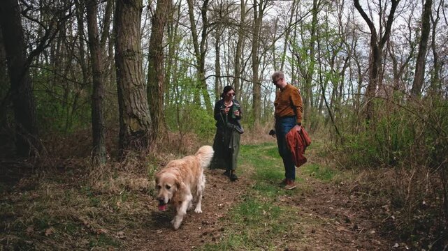 Two people walk along a path in the woods with a golden retriever dog. The trees surround them and the sky is gray as they enjoy their time together.