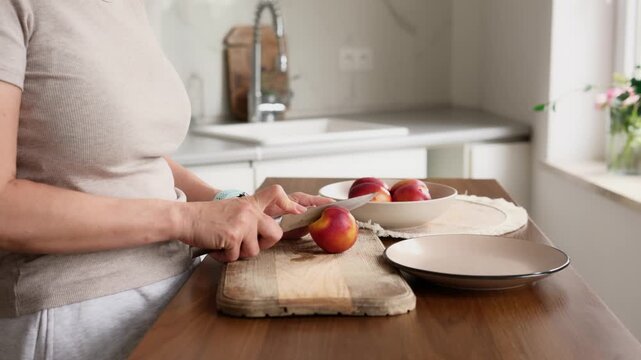 Woman carefully slices ripe peaches on a wooden board in a bright kitchen. Preparing fresh fruit for a snack.