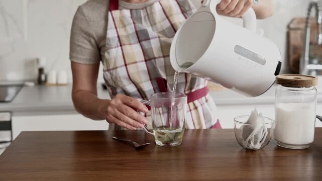 Woman in a plaid apron carefully pours hot water into a clear mug, making tea on a kitchen counter.