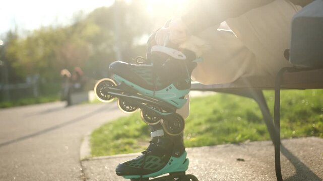 Girl secures teal and black inline skates while sitting on a park bench. Sunlight illuminates the paved path and green grass.