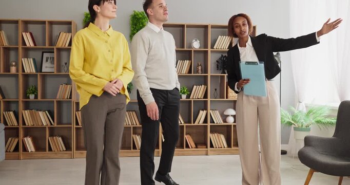 Agent showing couple buyers apartment property viewing. Professional agent gestures with a clipboard as clients review light, space and layout. Real estate consultation and viewing concept.