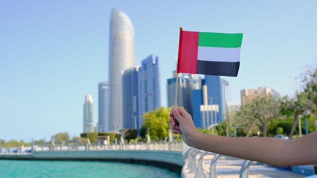 Female hand holding small UAE flag against Abu Dhabi skyline. Symbol of national pride and modernity with iconic skyscrapers in the background showcasing urban elegance and progress
