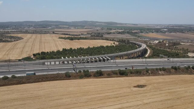 Aerial drone footage from high altitude circling a major highway, revealing a central traffic junction with multiple exits and bridges, surrounded by wheat fields, a commercial area, and a distant cit