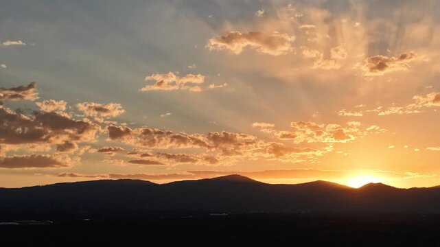 4K UHD drone timelapse: sweeping sky scale captures vivid golden sunbeams fanning through scattered cloud formations. Dynamic atmospheric transition highlights light rays over dark mountain ridges.