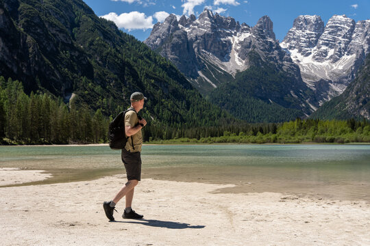 Male tourist with backpack walking on the sandy beach of Lake Landro and looking at majestic rocky mountain peaks, solo active hiking and summer wilderness exploration concept