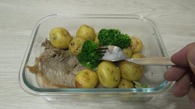 Cooked meat, baby potatoes and broccoli sit in a rectangular dish as a hand lifts a broccoli floret with a fork, showing homemade meal prep and portion control.
