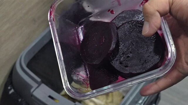 Unused cooked beet slices are tipped from a plastic container into a kitchen trash bin, showing everyday food waste and disposal habits.