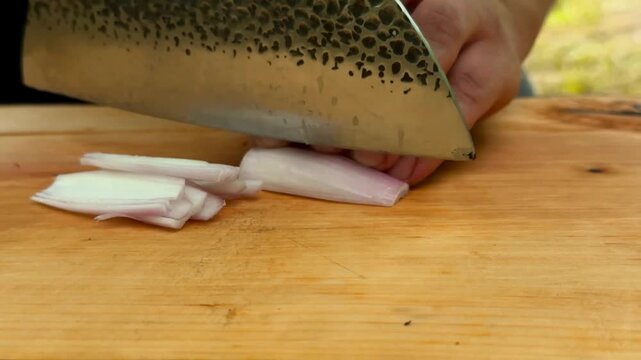 A person uses a sharp knife to carefully dice a fresh shallot on a wooden surface with a natural outdoor background which enhances the culinary preparation process for flavorful cooking recipes