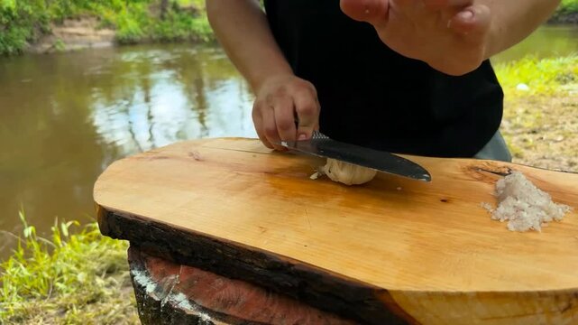 A person uses a sharp knife to peel and mince fresh garlic cloves on a rustic wooden surface with a river backdrop which assists in preparing flavorful ingredients for various kitchen recipes
