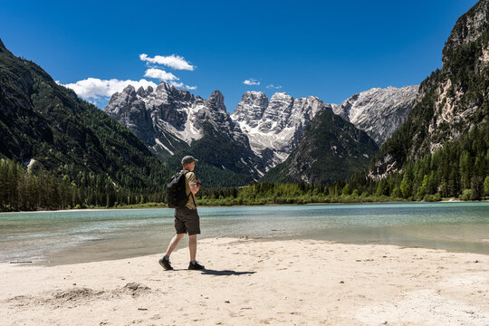 Male tourist with backpack walking on the sandy beach of Lake Landro and looking at majestic rocky mountain peaks, solo active hiking and summer wilderness exploration concept