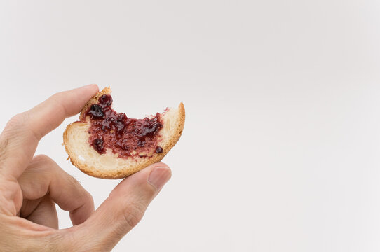 Primo piano di mano che regge una fetta di pane addentata con marmellata su uno sfondo bianco