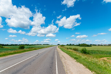 Fototapeta premium Open rural road in summer with lush green meadows and bright sky. Scenic asphalt road winding through green rural landscape under a blue cloudy sky.
