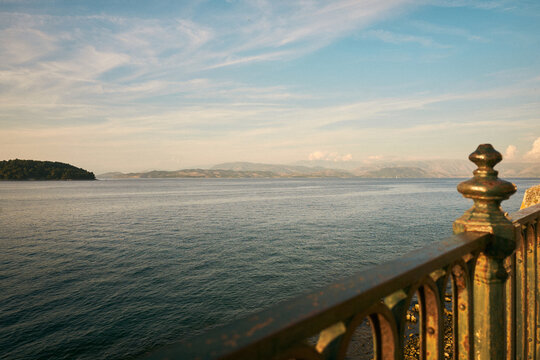 Entspannter Ausblick &uuml;ber das Meer auf eine ferne Berglandschaft, eingerahmt von einem historischen Metallgel&auml;nder an einer Uferpromenade auf Korfu.