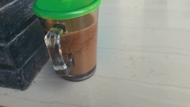 A clear plastic mug with a green lid containing a brown liquid sits on a white surface next to a stack of dark gray bricks