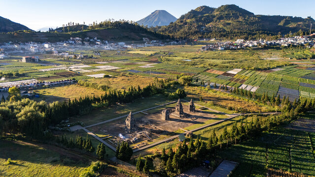 Aerial view of the historic Arjuna Temple complex surrounded by lush agricultural fields and mountains in Dieng Kulon, Jawa Tengah, Indonesia.