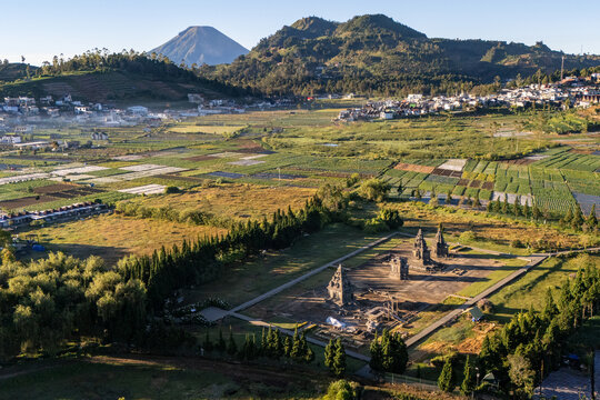 Aerial view of the Arjuna Temple complex surrounded by agricultural fields, lush greenery, and distant mountains in Dieng Kulon, Jawa Tengah, Indonesia.