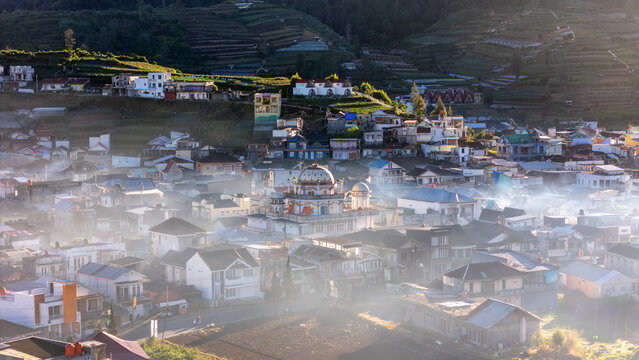 Aerial view of the village of Dieng Kulon with a central mosque, terraced hillsides, and morning mist rising between houses. Dieng Kulon, Jawa Tengah, Indonesia.