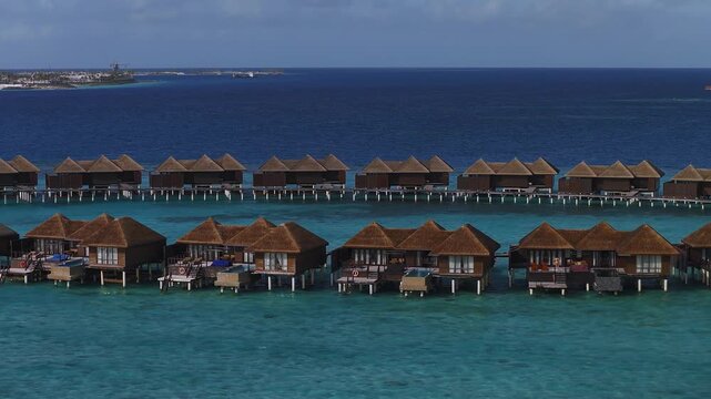 Aerial daytime pan shows Maldivian resort with two rows of thatched villas on stilts, wooden walkways, private decks, plunge pools, and boats near the distant island.