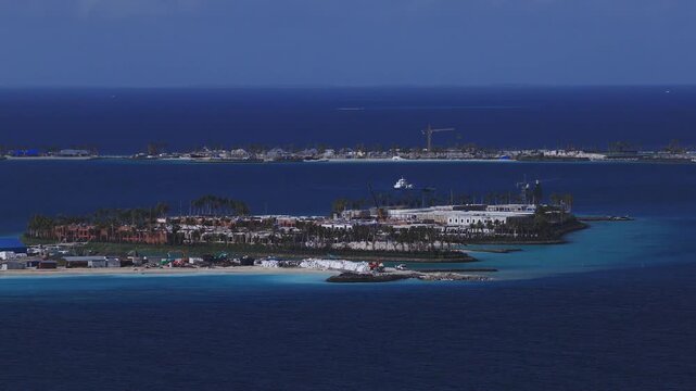 Aerial view of a Maldives resort island with white sand, turquoise lagoons, palms, low rise buildings, cranes, heavy machinery, and a workboat in bright daylight.