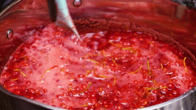 Boiling redcurrant berries in a pot to extract fresh juice for homemade jam. A chef gently stirs the hot mixture with a spatula. The bright red mass bubbles during traditional cooking.