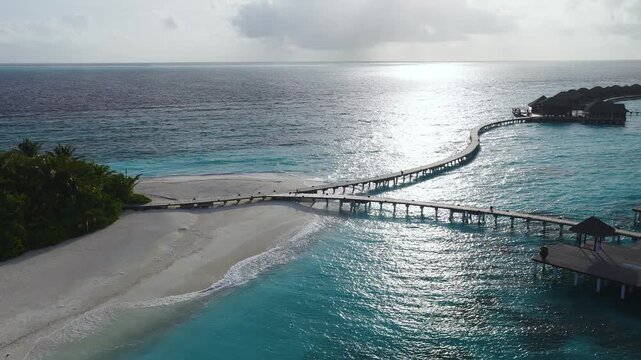 Aerial Maldives island resort shows curving wooden jetties to overwater villas, white sand, palm fringe, Indian Ocean, late afternoon sun path, tiny figures on piers.