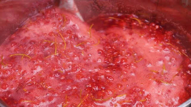 Boiling redcurrant berries in a pot to extract fresh juice for homemade jam. A chef gently stirs the hot mixture with a spatula. The bright red mass bubbles during traditional cooking.