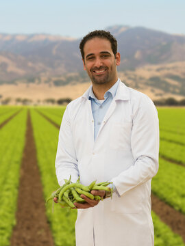 Smiling Middle Eastern agricultural scientist in white lab coat holding fresh fava beans in a green California farm field ideal for sustainable