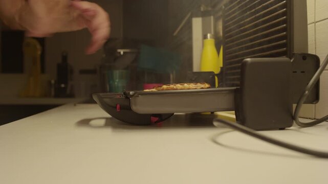 Burning toast smoking inside a sandwich maker at night. Man opens the toaster in a dark kitchen, releasing heavy smoke from the ruined burned food.