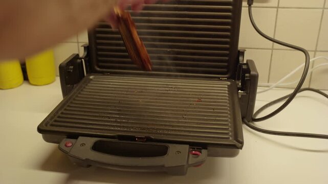 Thick smoke rises as a man's hand lifts the lid of a kitchen sandwich maker at night, revealing a completely burned toast. A dangerous midnight cooking accident.