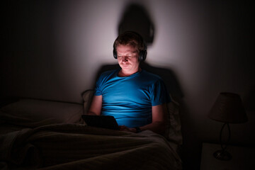 Obraz premium Man relaxing in bed at night using tablet and headphones