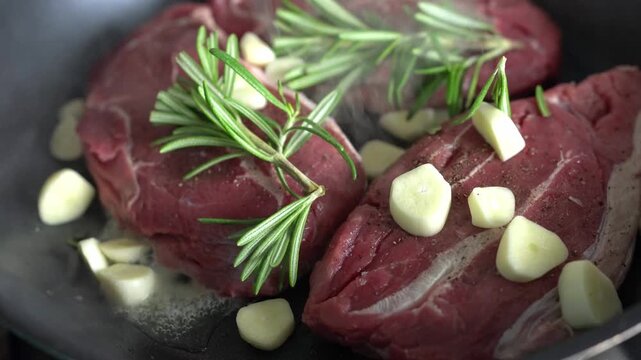 Close-up of steaks being cooked in a frying pan. The steaks are being fried in a pan of sizzling oil with garlic and a sprig of rosemary.