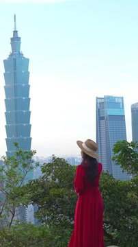 Woman in a flowing red dress stands on a scenic hilltop overlooking a modern city skyline. Travel, lifestyle, freedom, tourism, urban landscape and outdoor adventure concept.