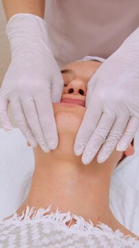Professional masseuse in white gloves performing a relaxing intraoral buccal massage on a young woman's face. Vertical video
