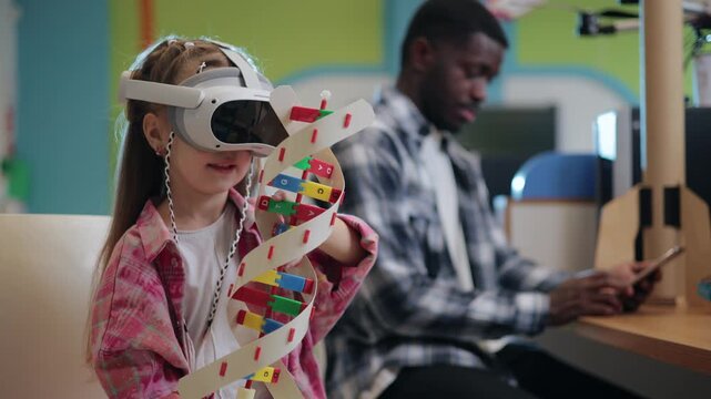 Kid girl with virtual reality headset exploring colorful dna model in classroom learning area with seated African teacher adult in background.